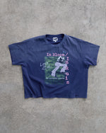 A faded navy blue cropped t-shirt with a distressed graphic print, laid flat on a textured gray concrete surface. The graphic features a green and white illustration of a person with pink text reading 'In Bloom/In Ruin' and 'Lost in Allure'.